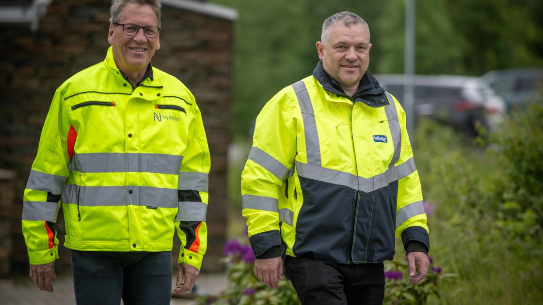 To menn i gule arbeidsjakker stående utendørs, omringet av grønn vegetasjon.