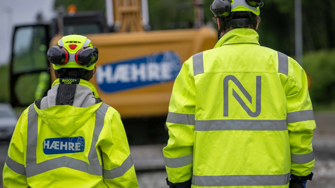 To personer i gule arbeidsjakker med logoene til Hæhre og Nye Veier, foran en anleggsmaskin.