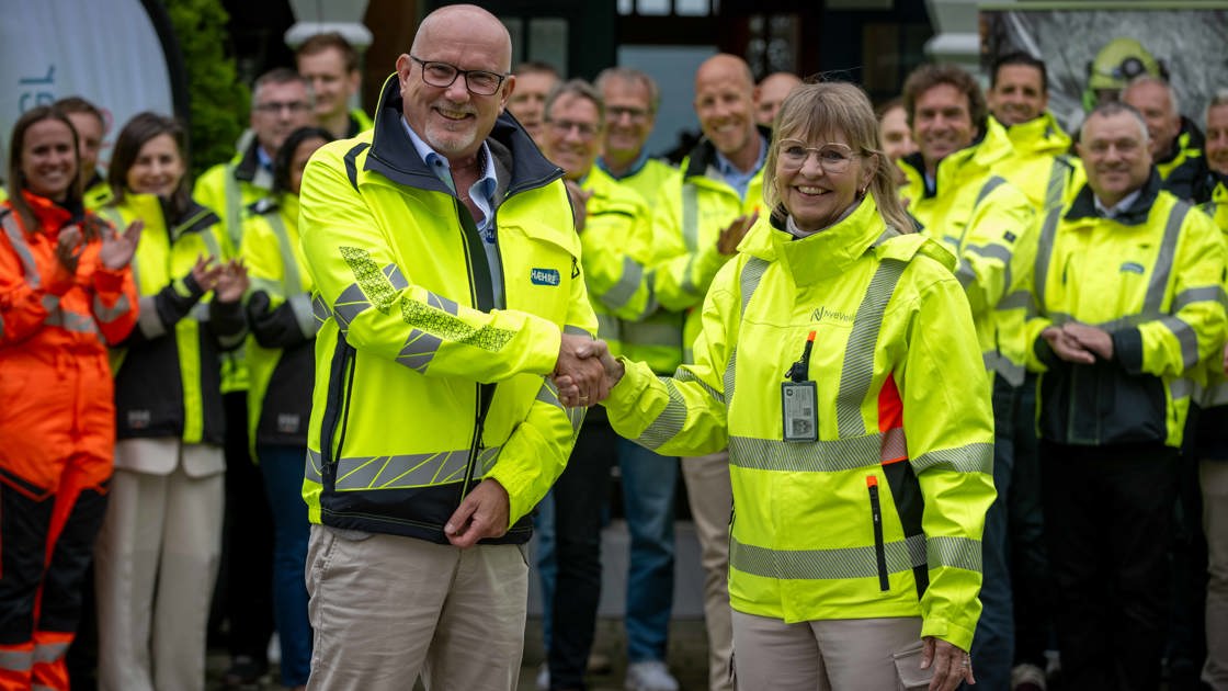 To personer i gule refleksjakker håndhilser foran en gruppe mennesker.